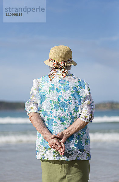 Rückansicht der älteren Frau mit Strohhut am Strand mit Blick in die Ferne