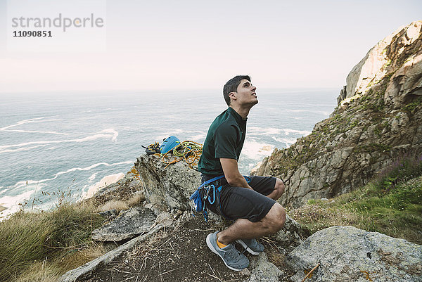 Kletterer sitzt auf einem Felsen an der Küste und schaut nach oben.