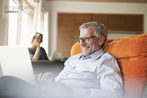 Lächelnder Mann mit Laptop im Sessel mit Frau im Hintergrund