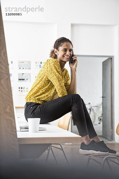 Lächelnde Frau auf dem Schreibtisch sitzend mit dem Handy