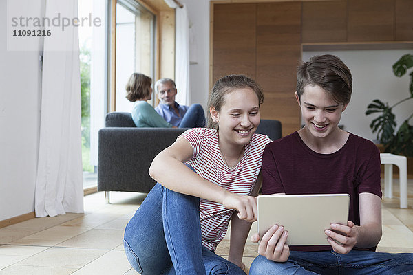 Lächelnde Teenager-Geschwister teilen sich die Tablette im Wohnzimmer mit den Eltern im Hintergrund.
