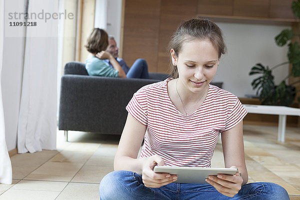 Lächelndes Mädchen mit Tablette im Wohnzimmer mit Eltern im Hintergrund