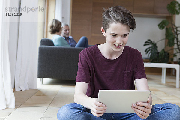 Lächelnder Teenager mit Tablette im Wohnzimmer mit Eltern im Hintergrund