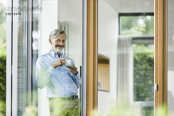 Lächelnder Mann mit Tasse Kaffee in seinem Haus und Blick durchs Fenster