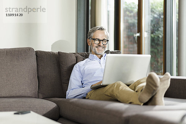 Lächelnder Mann sitzt auf der Couch in seinem Wohnzimmer mit Laptop