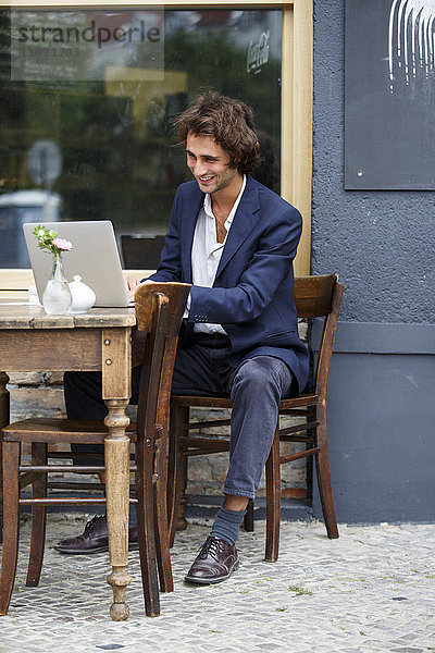 Lächelnder junger Geschäftsmann bei der Arbeit mit dem Laptop im Straßencafé
