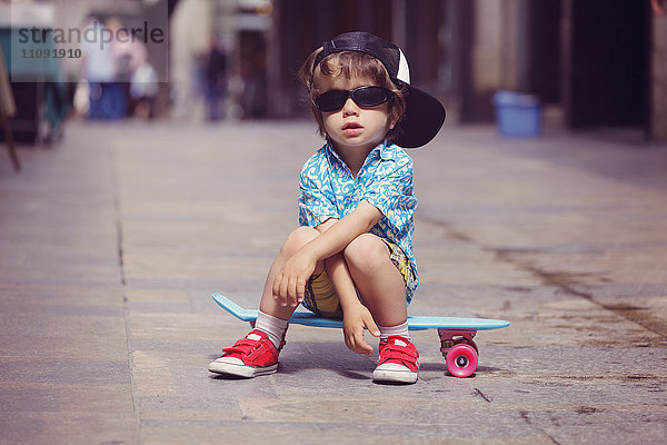 Porträt des kleinen Jungen auf dem Skateboard mit übergroßer Sonnenbrille und Basecap