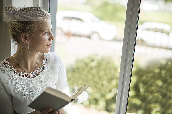 Junge Frau liest ein Buch am Fenster und schaut hinaus.