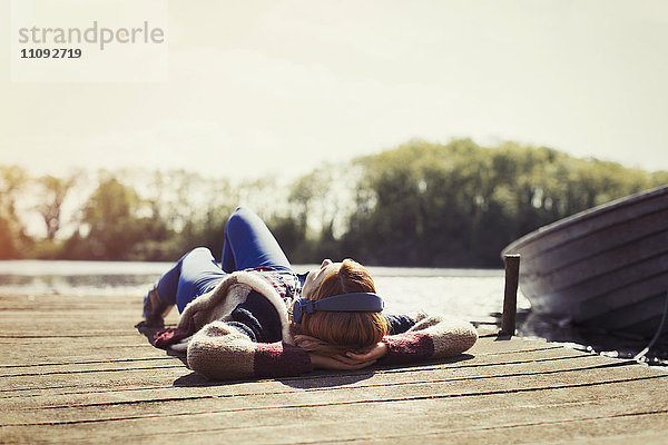 Frau entspannt auf dem Dock liegend Musik hören mit Kopfhörern am sonnigen Seeufer