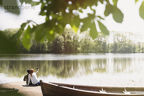 Paar mit Fotohandy  das Selfie am sonnigen Ufer des Sees neben dem Kanu mitnimmt.