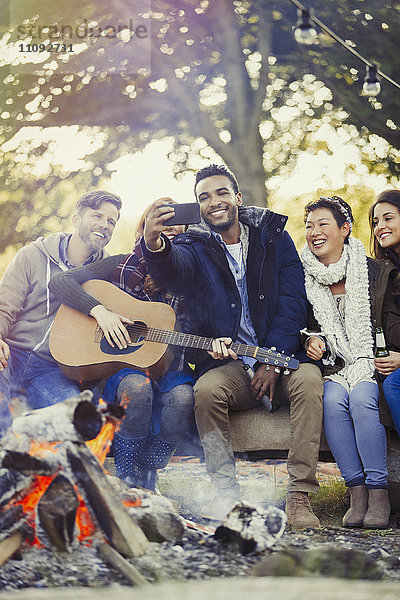 Freunde mit Gitarre nehmen Selfie mit Kamerahandy am Lagerfeuer