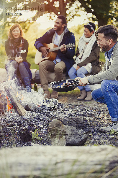 Freunde spielen Gitarre und kochen Hot Dogs am Lagerfeuer