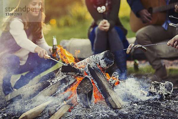 Freunde kochen Hot Dogs und trinken Bier am Lagerfeuer