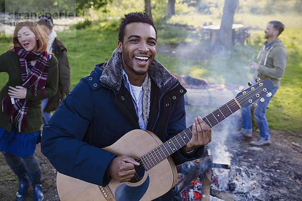 Porträt eines lächelnden Mannes  der mit Freunden auf dem Campingplatz Gitarre spielt.