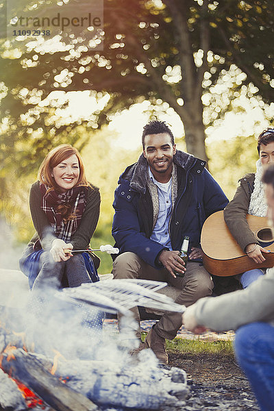 Lächelnde Freunde braten Marshmallows und trinken Bier am Lagerfeuer.