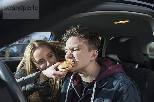 Junges Paar sitzt in einem Auto und sie füttert ihn mit einem Burger