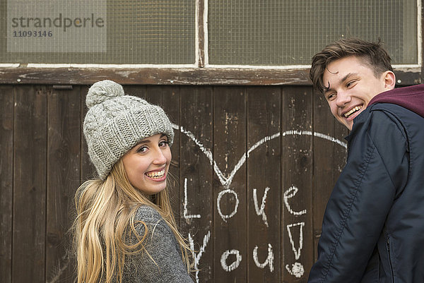 Junges Paar  das an einer Wand steht  auf der ein Herz gezeichnet und Liebe dich auf eine Holzwand geschrieben ist