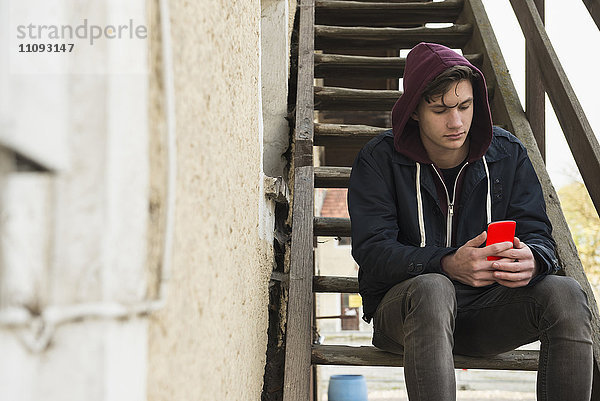 Junger Mann schreibt Textnachrichten auf seinem Smartphone und sitzt auf einer Holztreppe