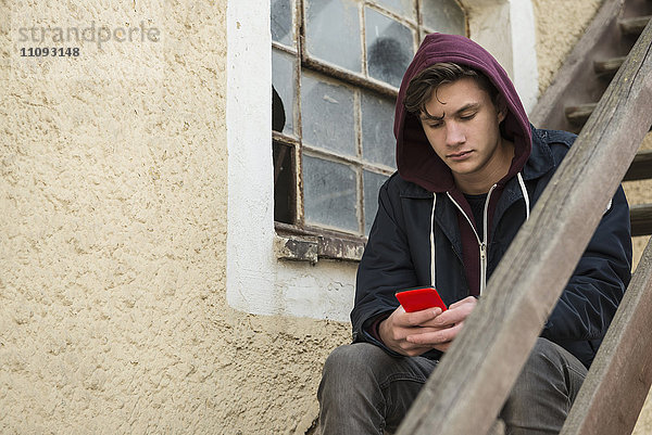 Junger Mann schreibt Textnachrichten auf seinem Smartphone und sitzt auf einer Holztreppe