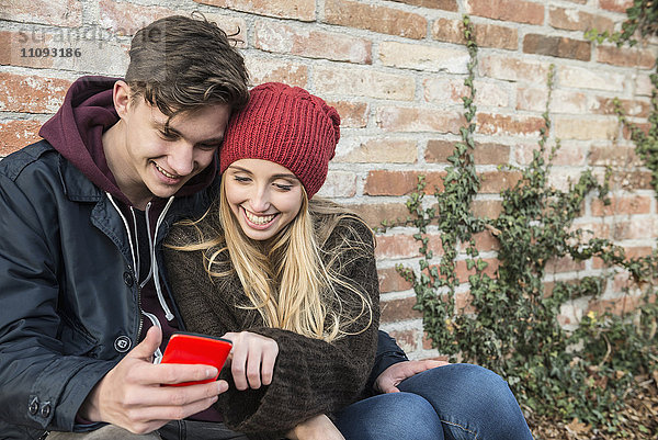 Junges Paar lächelt und benutzt ein Smartphone an einer Mauer