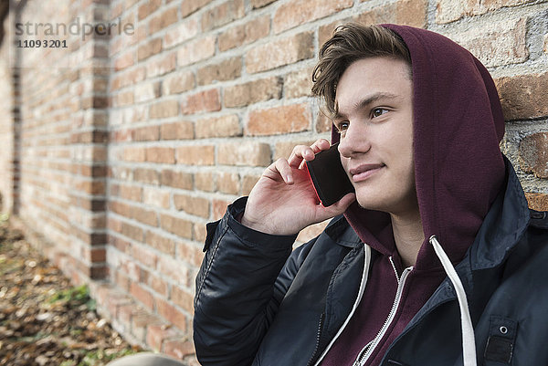 Junger Mann spricht mit einem Handy und lehnt sich an eine Backsteinmauer