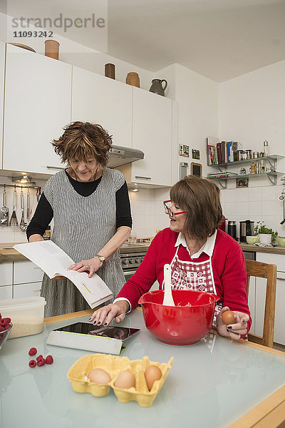 Zwei ältere Frauen vergleichen Rezepte auf einem digitalen Tablet und in einem Kochbuch