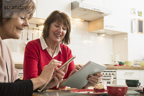 Zwei ältere Frauen benutzen ein Smartphone und ein digitales Tablet in der Küche