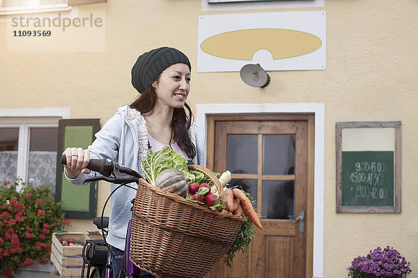 Mittlere erwachsene Frau mit Fahrrad und Gemüse vor einem Naturkostladen stehend