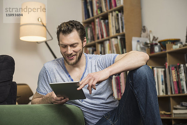 Mittlerer erwachsener Mann mit digitalem Tablet im Wohnzimmer und lächelnd  München  Bayern  Deutschland