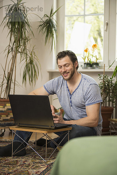 Mittlerer erwachsener Mann  der im Wohnzimmer an einem Laptop arbeitet und lächelt  München  Bayern  Deutschland