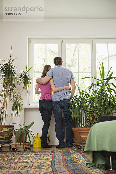 Rückansicht eines Paares  das im Wohnzimmer steht und durch ein Fenster schaut  München  Bayern  Deutschland