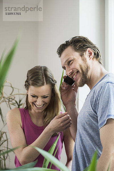 Mittlerer erwachsener Mann  der im Wohnzimmer mit einem Mobiltelefon spricht und lächelt  München  Bayern  Deutschland
