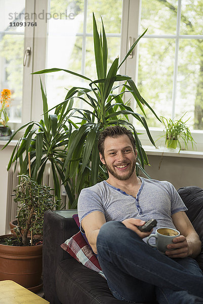 Mittlerer erwachsener Mann sieht fern und trinkt Kaffee im Wohnzimmer  München  Bayern  Deutschland