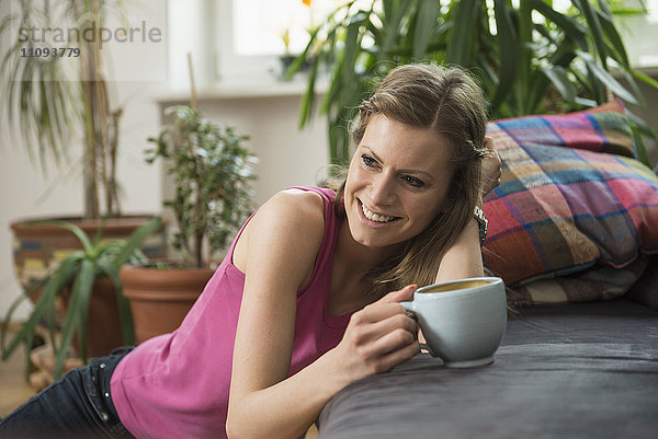 Junge Frau trinkt Kaffee im Wohnzimmer und lächelt  München  Bayern  Deutschland