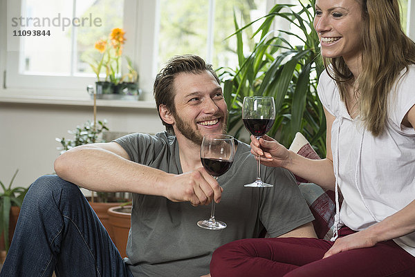 Paar trinkt Rotwein im Wohnzimmer und lächelt  München  Bayern  Deutschland