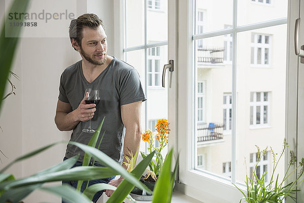 Mittlerer erwachsener Mann schaut durch ein Fenster im Wohnzimmer und trinkt Rotwein  München  Bayern  Deutschland