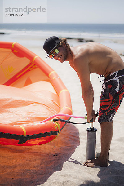 Junger Mann pumpt Luft in ein Kiteboard am Strand