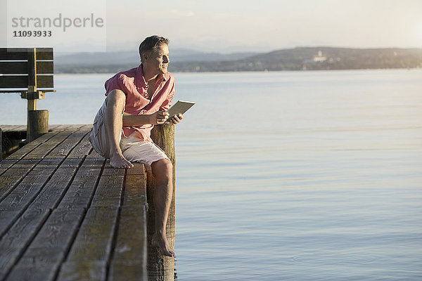 Älterer Mann mit digitalem Tablet auf einem Steg und Blick über einen See  Bayern  Deutschland