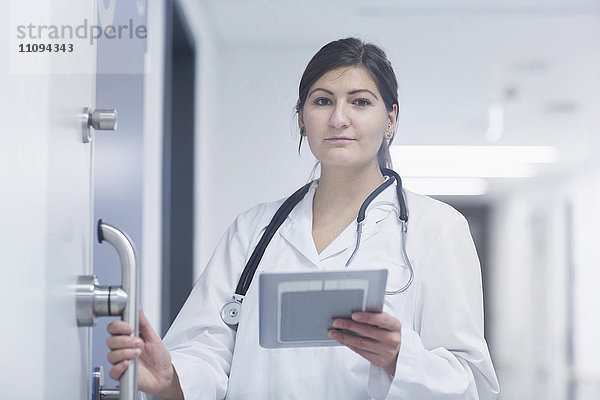 Porträt einer jungen Ärztin  die in einem Krankenhauskorridor ein digitales Tablet benutzt
