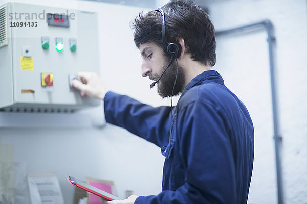 Junger männlicher Ingenieur mit Headset  der mit Hilfe eines digitalen Tablets in einer Industrieanlage die Steuertafel aktualisiert