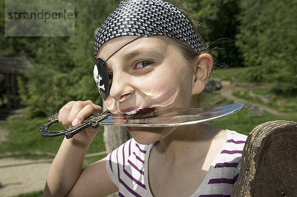 Porträt eines als Pirat verkleideten Mädchens  das auf einem Spielplatz einen Säbel beißt  Bayern  Deutschland