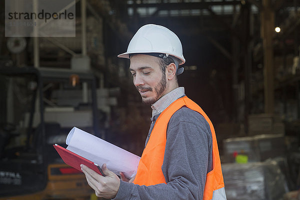 Junger männlicher Ingenieur hält einen Bauplan und benutzt ein digitales Tablet auf einer Baustelle