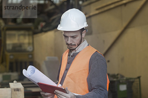 Junger männlicher Ingenieur hält einen Bauplan und benutzt ein digitales Tablet auf einer Baustelle