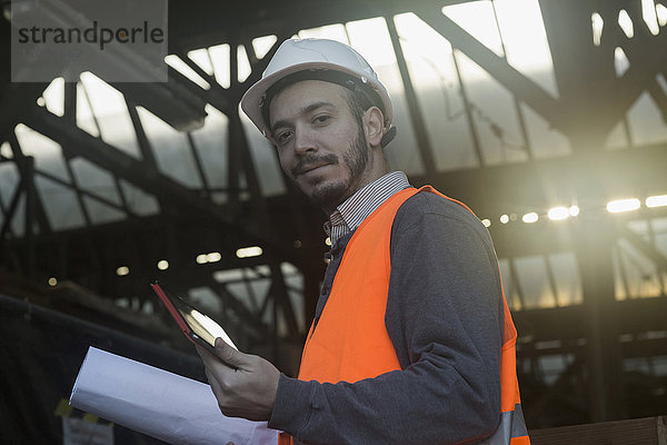 Porträt eines jungen männlichen Ingenieurs  der einen Bauplan hält und ein digitales Tablet auf einer Baustelle benutzt