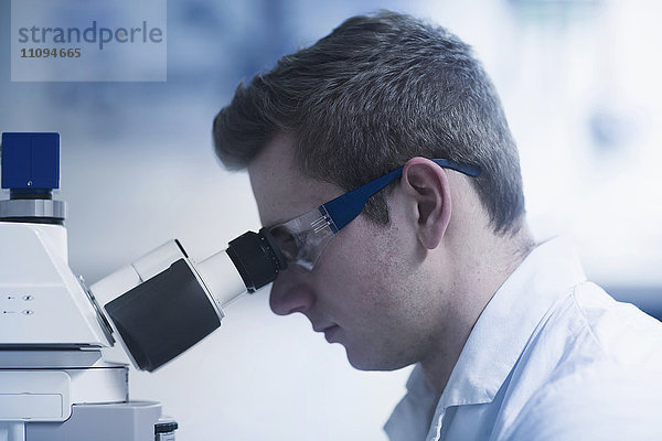 Junger männlicher Wissenschaftler  der durch ein Mikroskop in einem optischen Labor schaut