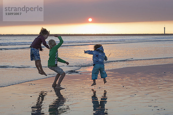 Drei Kinder springen bei Sonnenuntergang am Strand  Viana do Castelo  Region Norte  Portugal