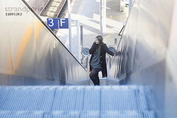 Junge Frau  die sich auf einer Rolltreppe bewegt und auf dem Bahnhof telefoniert  Freiburg im Breisgau  Baden-Württemberg  Deutschland
