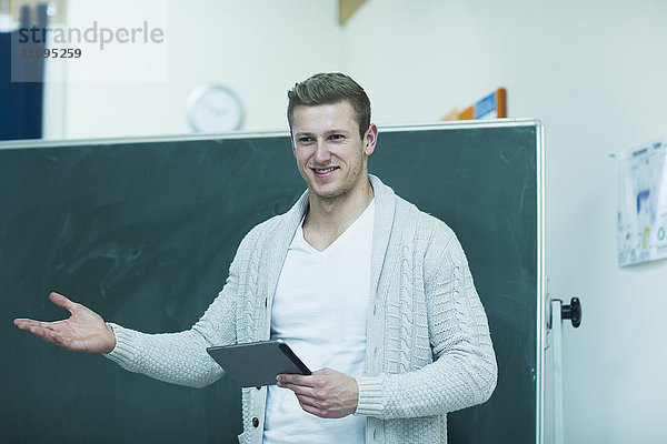 Junger männlicher Lehrer  der ein digitales Tablet hält und an der Tafel im Klassenzimmer erklärt  Freiburg im Breisgau  Baden-Württemberg  Deutschland