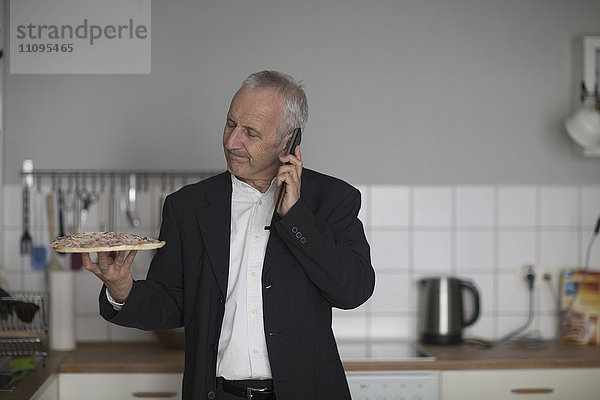 Älterer Geschäftsmann isst Pizza und telefoniert mit dem Handy in der Küche  Freiburg im Breisgau  Baden-Württemberg  Deutschland