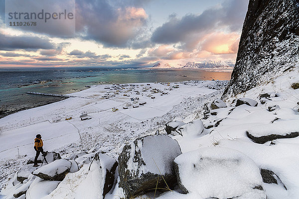 Fotograf auf dem Schnee bewundert das Fischerdorf unter einem bunten Himmel Eggum  Vestvagoy (Vest-Vagoy) Insel  Lofoten Inseln  Arktis  Norwegen  Skandinavien  Europa